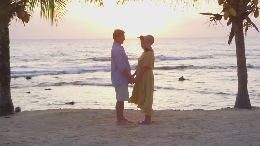 A man and woman standing on the beach facing each other holding hands