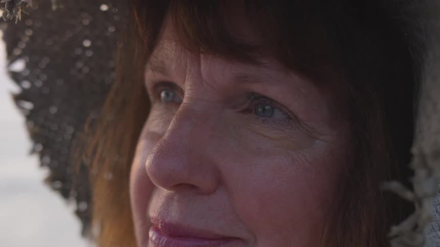 A woman with blue eyes and brown hair wears a straw hat while the sun shines upon her weathered face