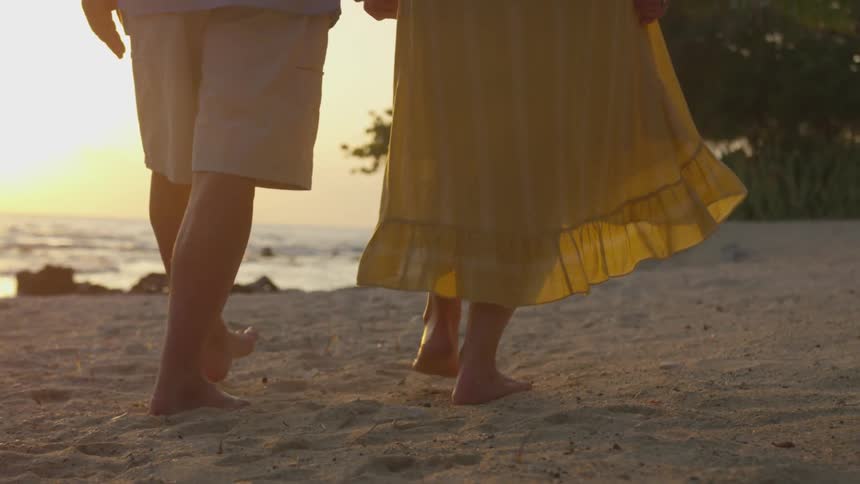 The bare feet and legs of a couple as they walk on the beach sand at sunset holding hands