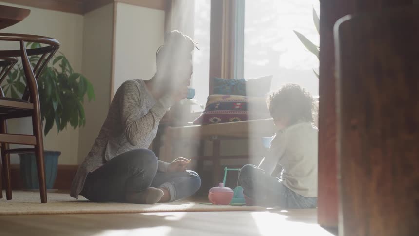 A young black woman sits on the floor and pretends to have a tea party with a young child