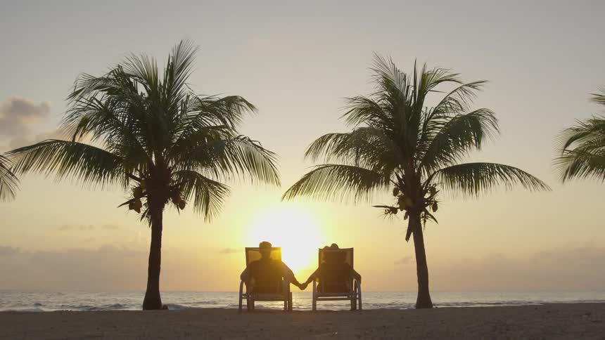 Two people holding hands while sitting in lounge chairs on the beach at sunset