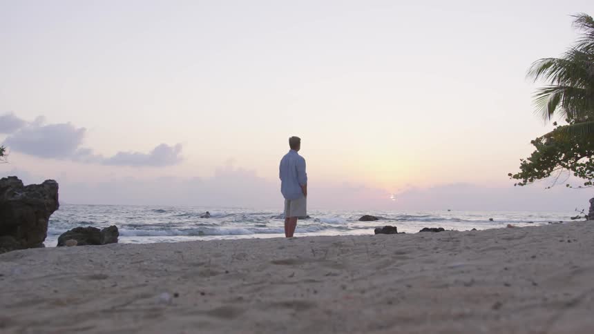 A man is standing alone in the sand on the beach watching the waves roll in while the sun is setting in the horizon
