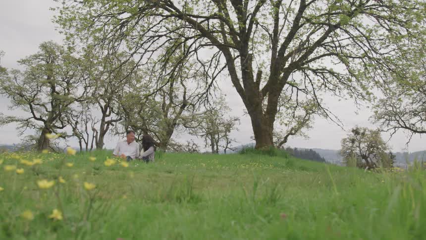 A man and a woman sit outdoors in a wide open grassy meadow as they converse