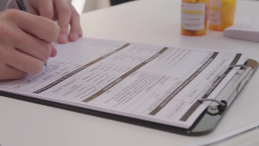 A pharmacist fills out a form on a desk which holds a stethoscope and pill bottles