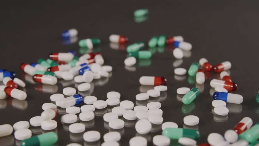 A large number of white and multicolored pills are dropped onto a shiny surface