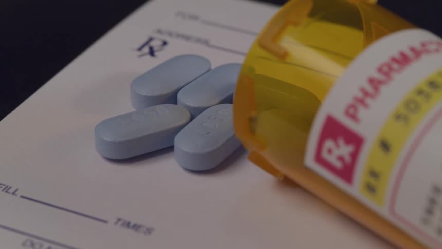 A bottle of blue prescription pills is open on its side and a few pills have spilled out on top of a pad of paper used to write prescriptions