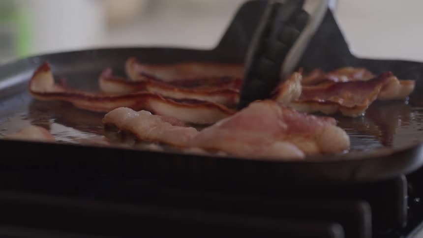 Bacon frying on griddle on stove top is turned