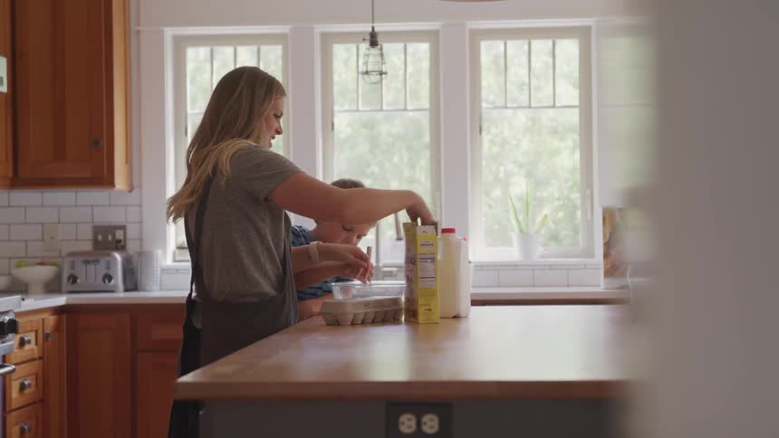 Mother smiles and pours flour into a mixing bowl as she and young son talk while preparing food together in the kitchen on a sunny afternoon