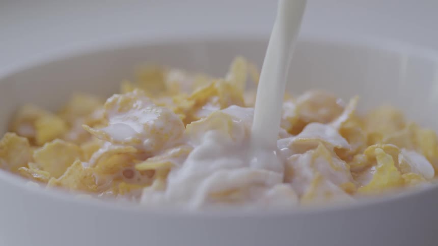 Milk pours onto a white bowl filled with corn flakes