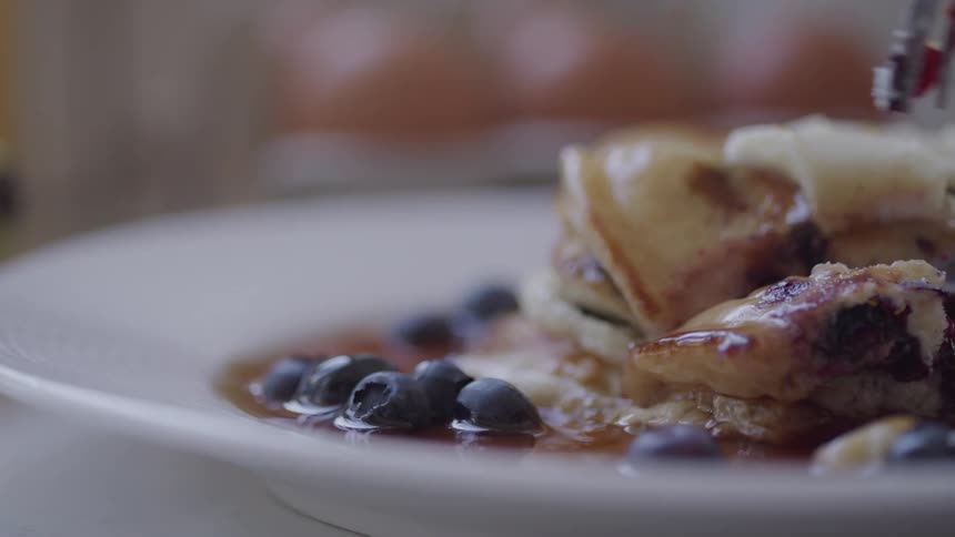 A knife cuts through blueberry pancakes and a wedge dripping with syrup is picked up with a fork
