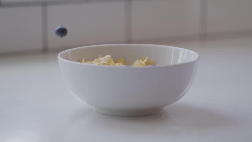 Blueberries are slowly dropped into a white bowl that contains cereal