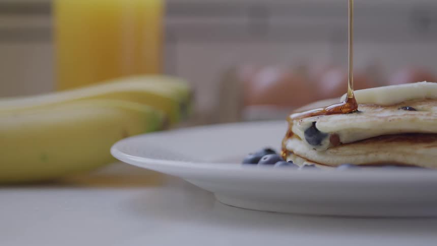 Syrup is slowly poured on blueberry pancakes on a white plate with bananas and juice nearby
