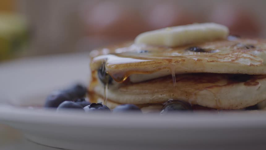 Syrup is poured onto and slowly drips off of a stack of blueberry pancakes on a plate