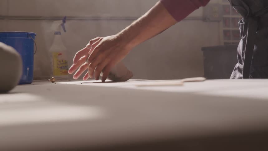 A man in a red shirt and black apron kneads some clay on a table