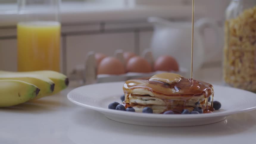Syrup pouring onto stack of pancakes with blueberries on a white plate next to bananas and a glass of orange juice
