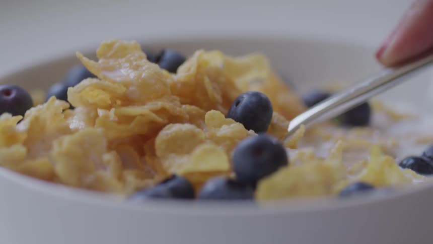 A person is using a spoon to scoop up some cereal topped with blueberries in a white bowl