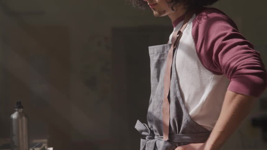 A male artist wearing an apron is looking intensely at something on a work table