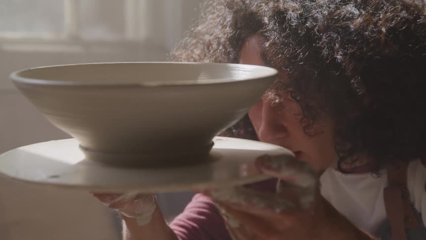A man holds a clay bowl on a potters wheel and examines it closely