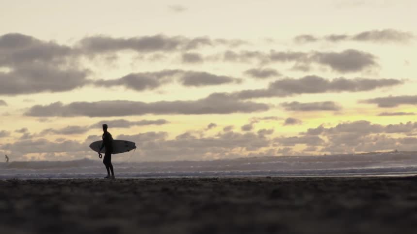 Man carrying a surfboard walking on the beach at sunset along the shoreline.