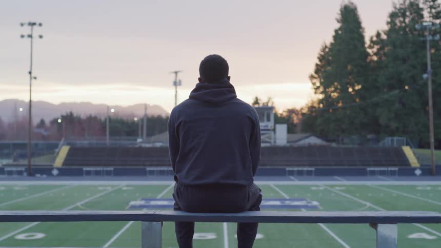 Man contemplating on a bench at the side of a football field