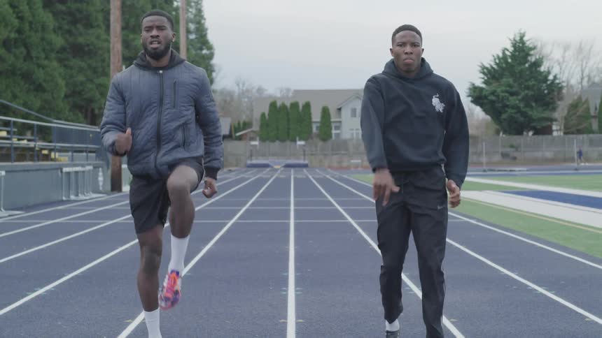 Two men wearing exercise outfits marching around race track and lifting their knees high