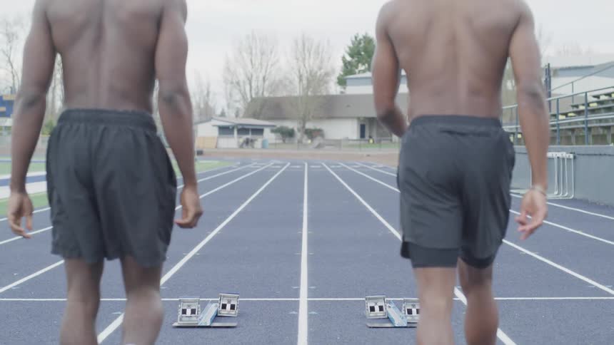 Two men who are not wearing shirts are each walking in a lane of a track towards a pair of starter blocks
