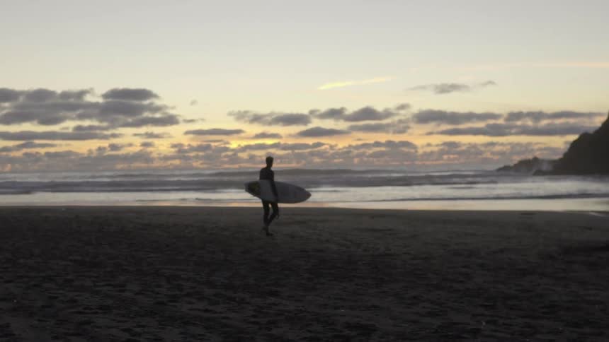 A surfer walks across the beach at sunset, carrying his board as he heads toward the waves.