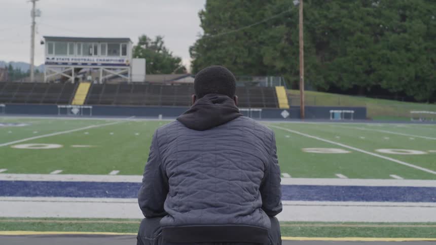 A man in a quilted coat sits near the sideline of a football field on the opposite side of the grand stands