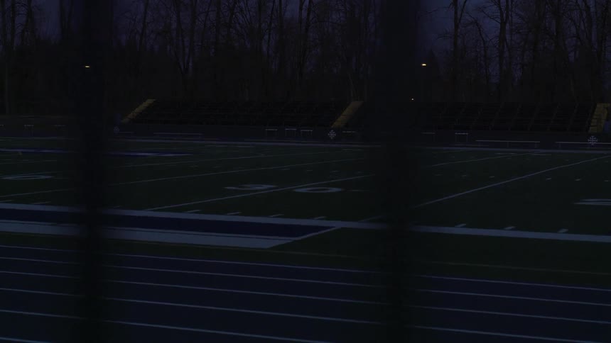 Through the darkness of night a football field can be seen that is lined by bare and leafless trees