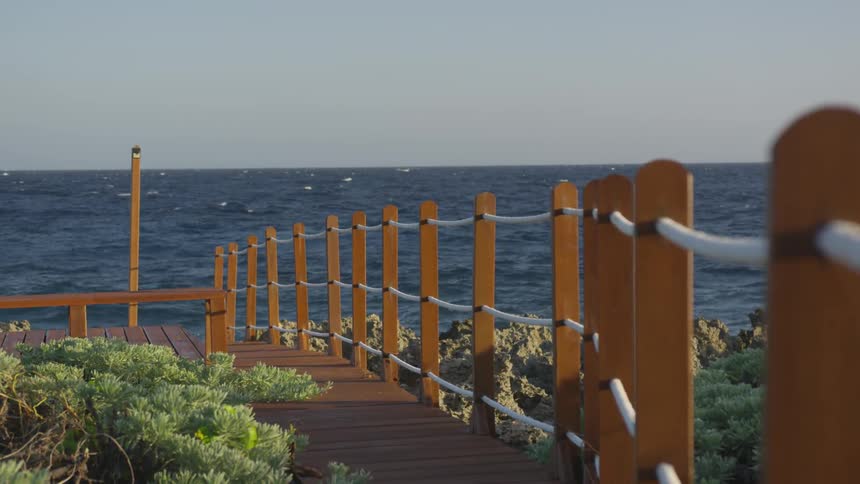 A wooden walkway that has a rope fence along it leading towards the ocean on a sunny day
