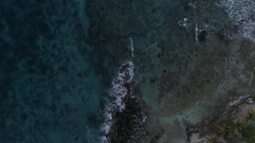 Ocean waves crash upon a rocky shoreline that is partially shielding some sandy beach in a tropical area