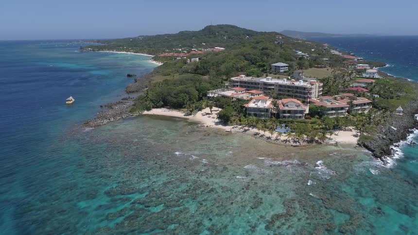 An island covered in trees and buildings is surrounded by clear blue water and a boat sails nearby