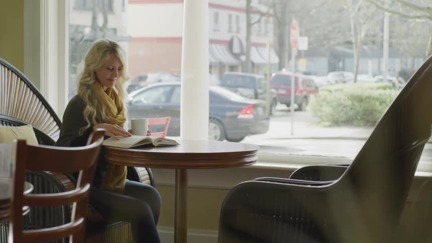 Blonde woman sits at the table and reads book while holding a cup in the cafeteria at daylight