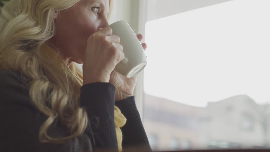 Elder blonde hair woman looks out the window and drinks out of a white mug indoors during daylight