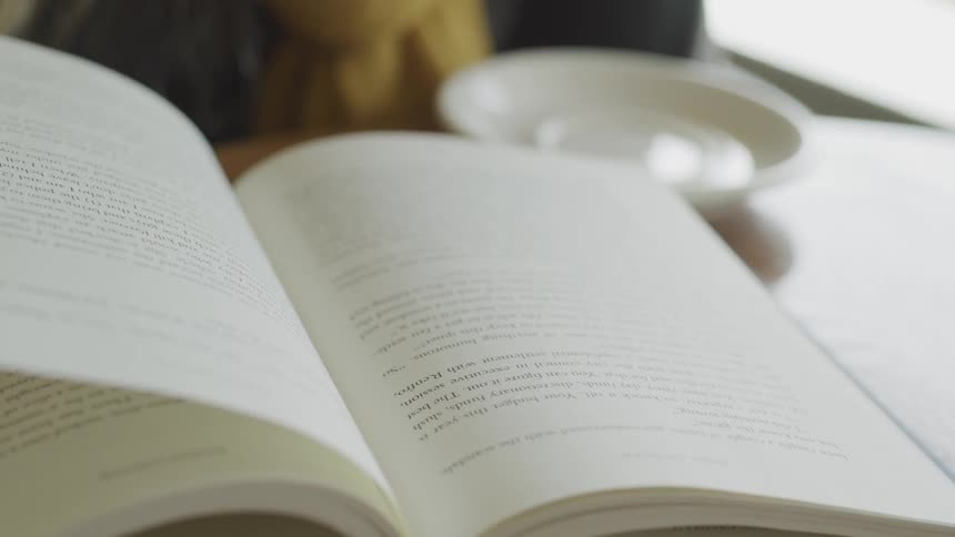 A person turning the pages of a book with one hand and holding a teacup with the other hand