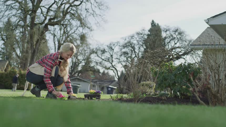 Woman is kneeling next to a flower bed in front of the house while digging a hole with a small shovel to plant flowers and a man is walking down the sidewalk behind her