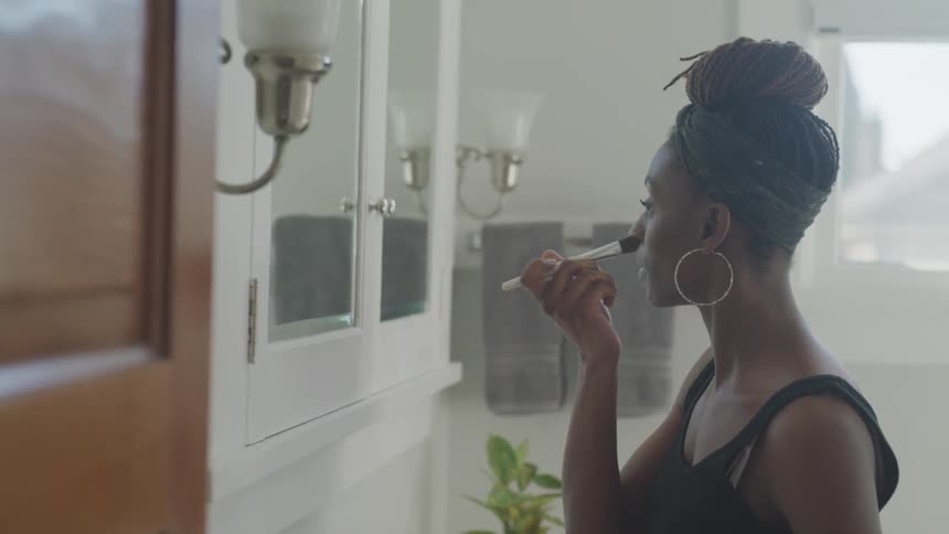 A woman in a black dress and large hoop earrings is applying makeup in front of her bathroom mirror