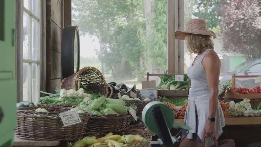 A blonde female in a sun hat is shopping for produce at an indoor farmers market