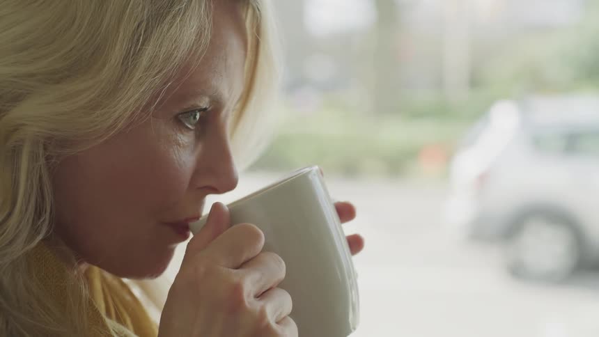 A woman smiles while sipping a hot drink from a white mug in front of a window