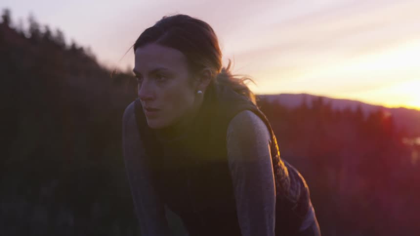 A woman in a black vest leans over to catch her breath in a forest at sunset