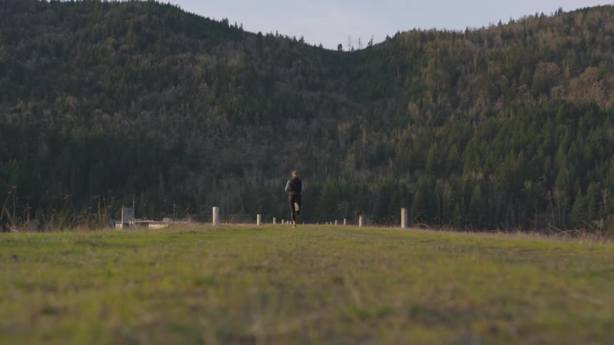 A woman runs through an open grassland toward a large tree lined mountain