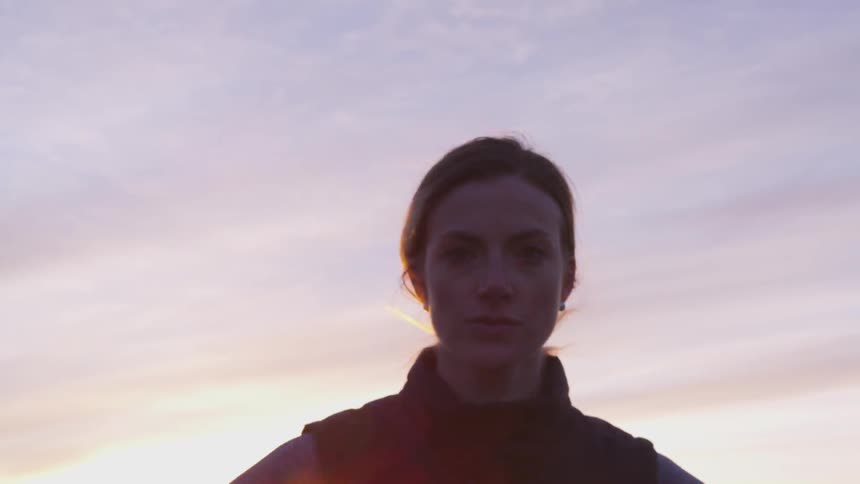 Woman wearing a vest standing outdoors with her hands on her hips as the sun goes down behind distant hills