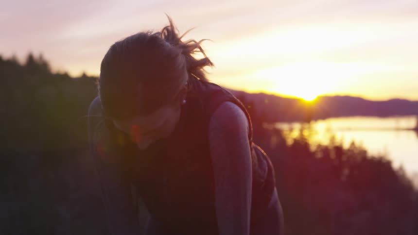 A woman bends over with hands on knees as she catches her breath standing on a hill at sunset