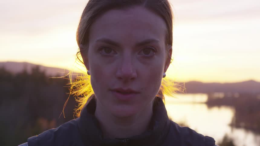 A woman with her hair tied back looks into the camera at sunset