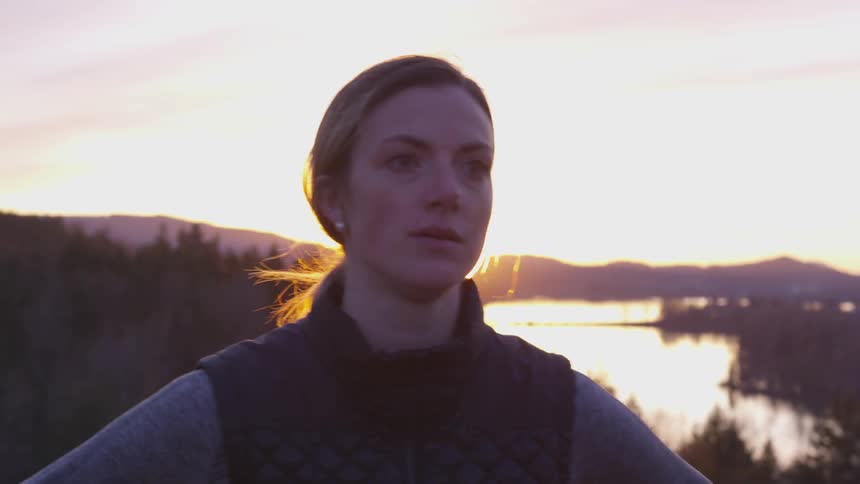 A woman breathes heavily while standing near a mountain lake at sunset
