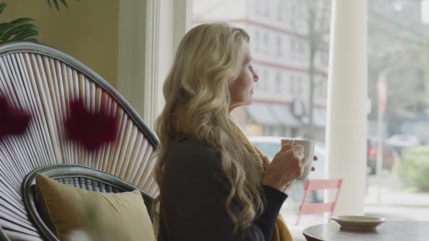 Woman sitting at the table holds mug and look outside the window in a cafe during daylight