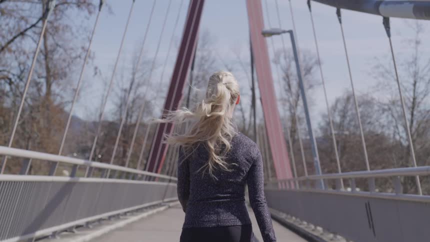 A womans blonde hair bounces as she speed walks across a pedestrian bridge