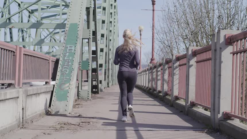 A woman with blonde hair jogs alone on the pedestrian path of a grey metallic bridge on a clear day