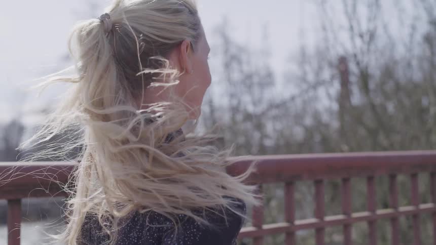 A white female with long blonde hair in a pony tail runs over a bridge with bare trees in the background