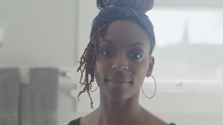 A black woman with a nose piercing and braids piled on top of her head turns slowly out of profile to face the camera